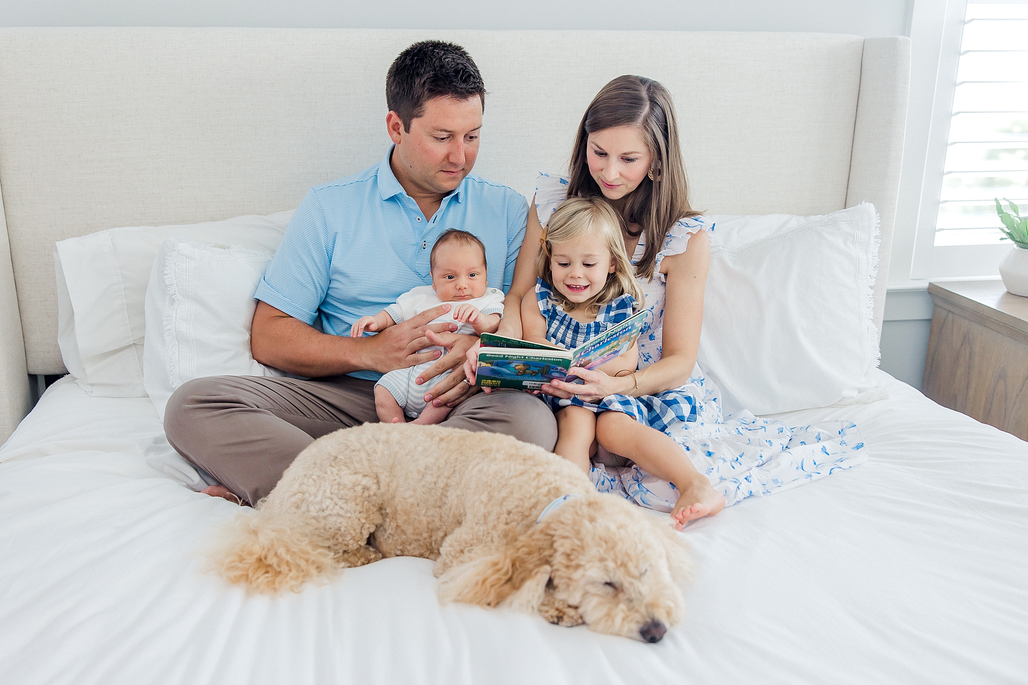 family of four snuggle and read a book on a bed with their dog nearby
