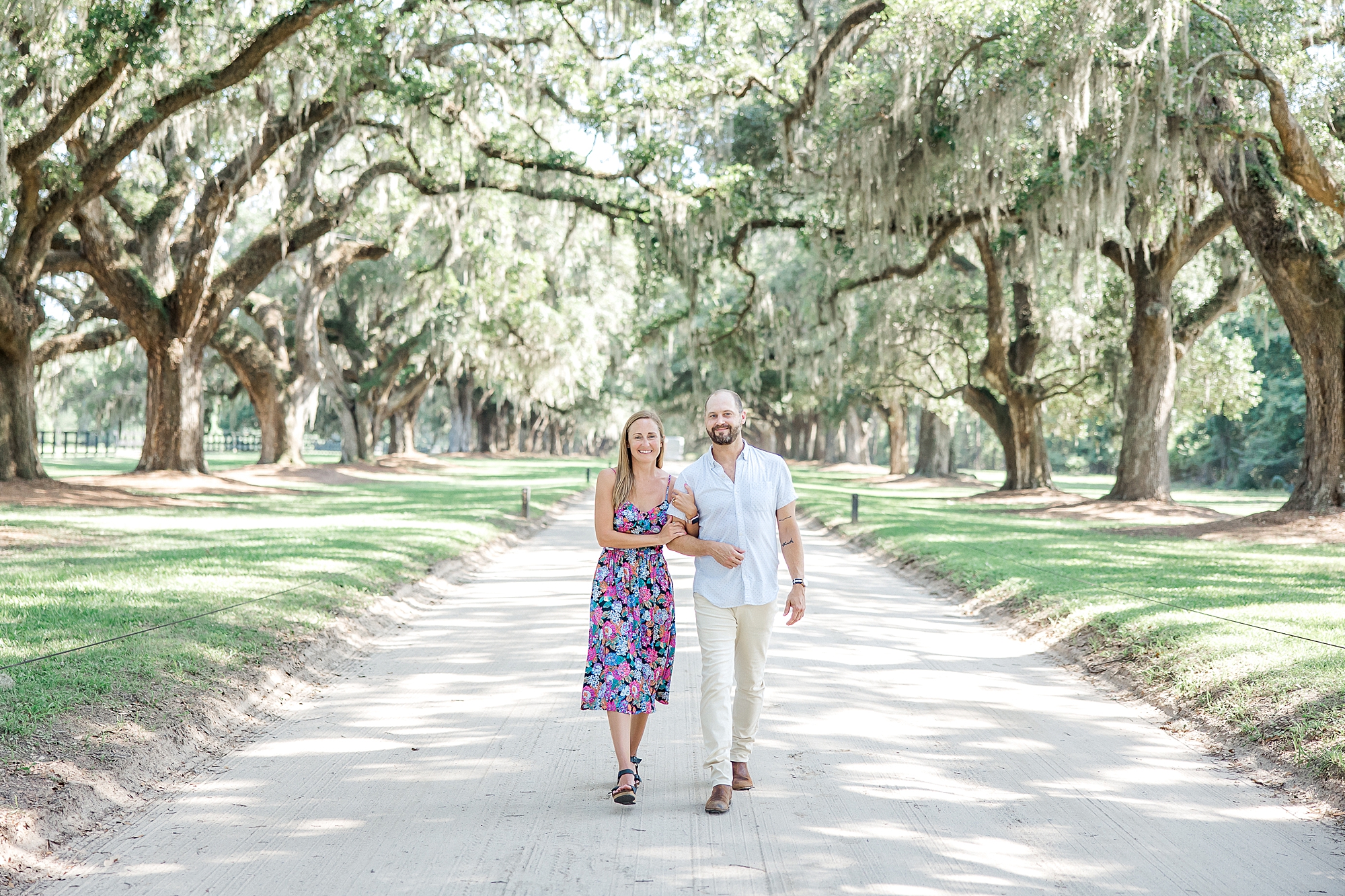 couple hold hands as they walk under oak trees covered in Spanish moss
