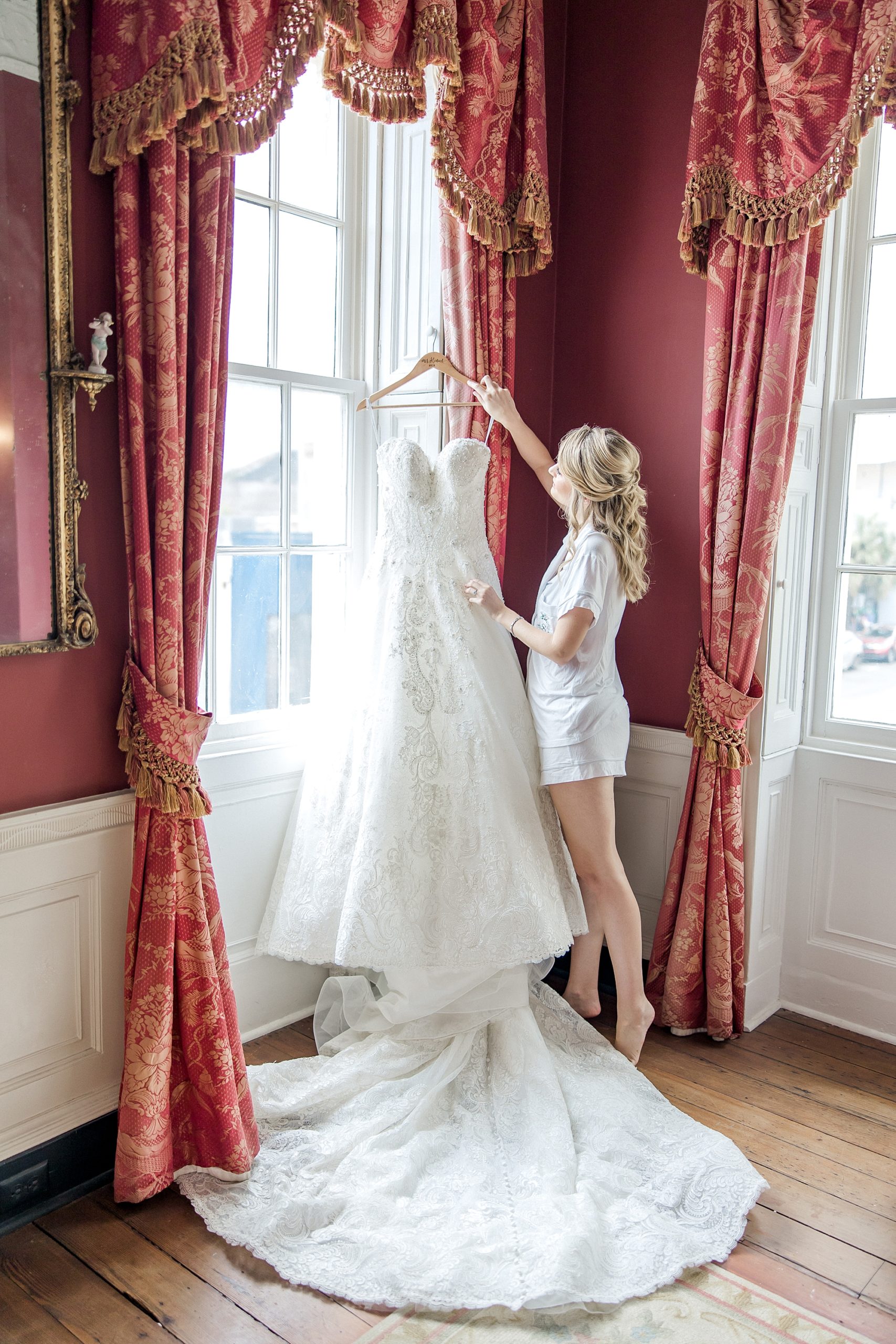 bride admires wedding dress hanging by the window