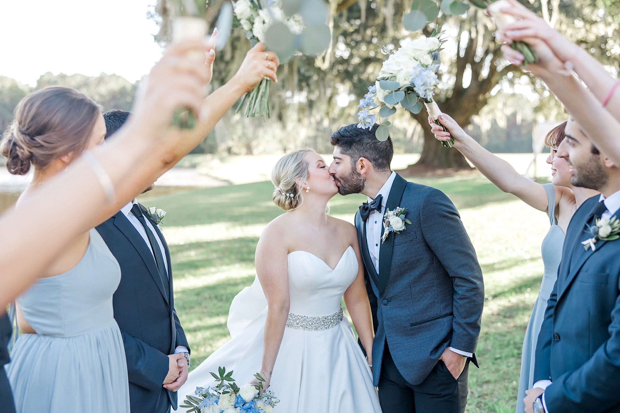 bride and groom kiss while wedding party lift up their arms around them